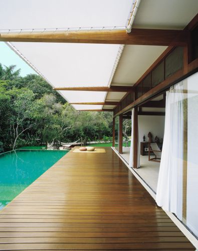 A varanda/deck que integra piscina e o living da casa é protegida pelo toldo de poliéster branco microperfurado. Ao fundo, o espaço para as redes pertence ao jardim e desfruta da proximidade da área de preservação ambiental