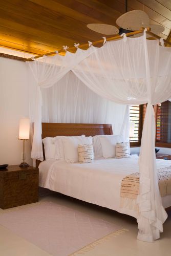 A ambientação da suíte máster acompanha o padrão do restante da casa: despojada e elegante. A cama com dossel de madeira e cortinado branco está em perfeita harmonia com o forro em réguas de madeira.