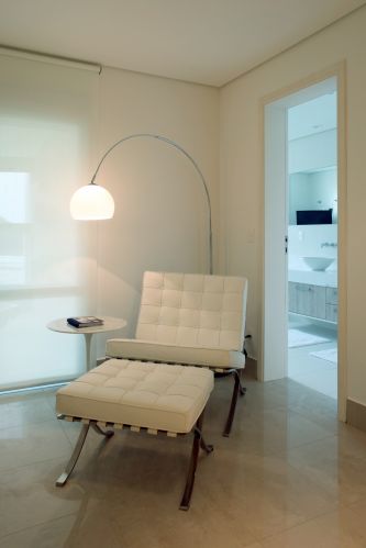 A luminária da R.R. Lustres ilumina a poltrona e pufe Brcelona, criando um canto de leitura. Na imagem ainda é possível vislumbrar o banheiro do casal, totalmente branco