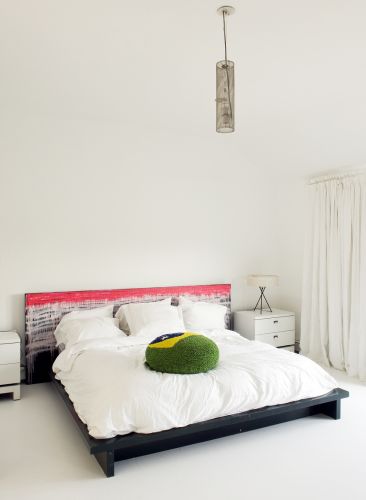No quarto do casal, a cama ganha destaque por suas cores fortes e estrutura próxima ao chão. Sobre a fofa roupa de cama branca, uma almofada reproduz a bandeira do Brasil