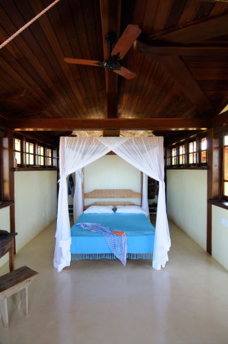 Com domínio de madeira em padrão mogno e branco, o quarto tem espaço amplo e foco principal na cama localizada no centro do ambiente. O dossel suaviza a rusticidade da madeira